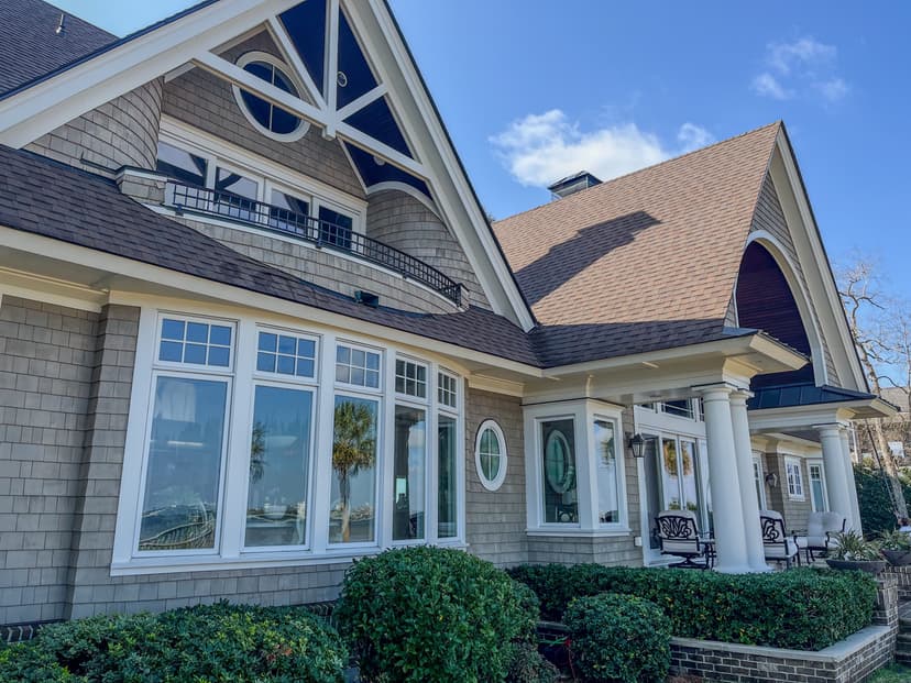 Elegant waterfront home with large windows, intricate roof design, and lush landscaping.